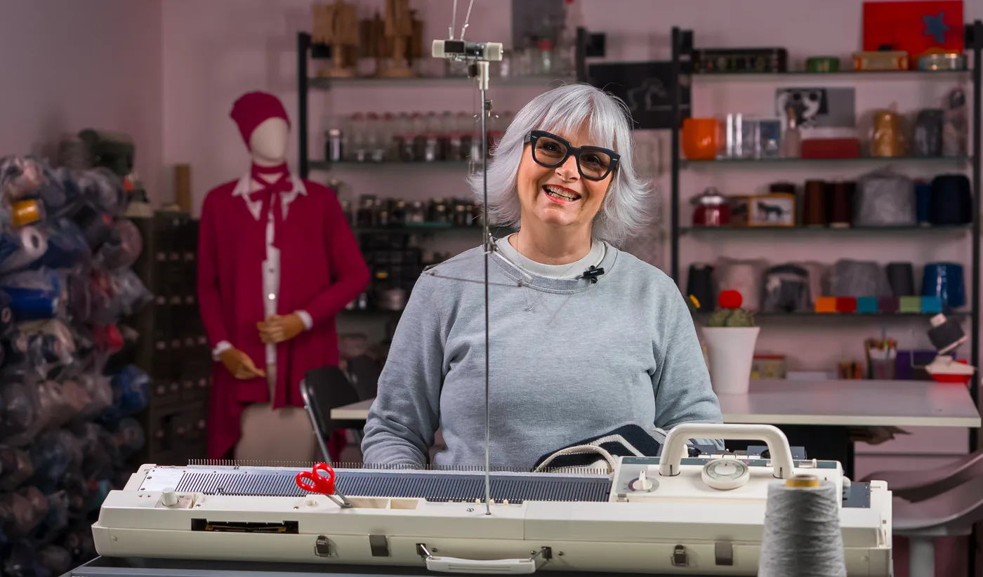 Donatella a una macchina da maglieria domestica nel suo studio, con una parete di filati alle spalle